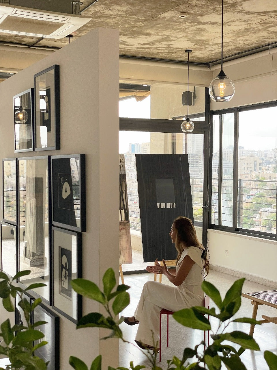 Rita Rawashdeh seated in a sunlit studio surrounded by framed artwork, talking with city views in the background.