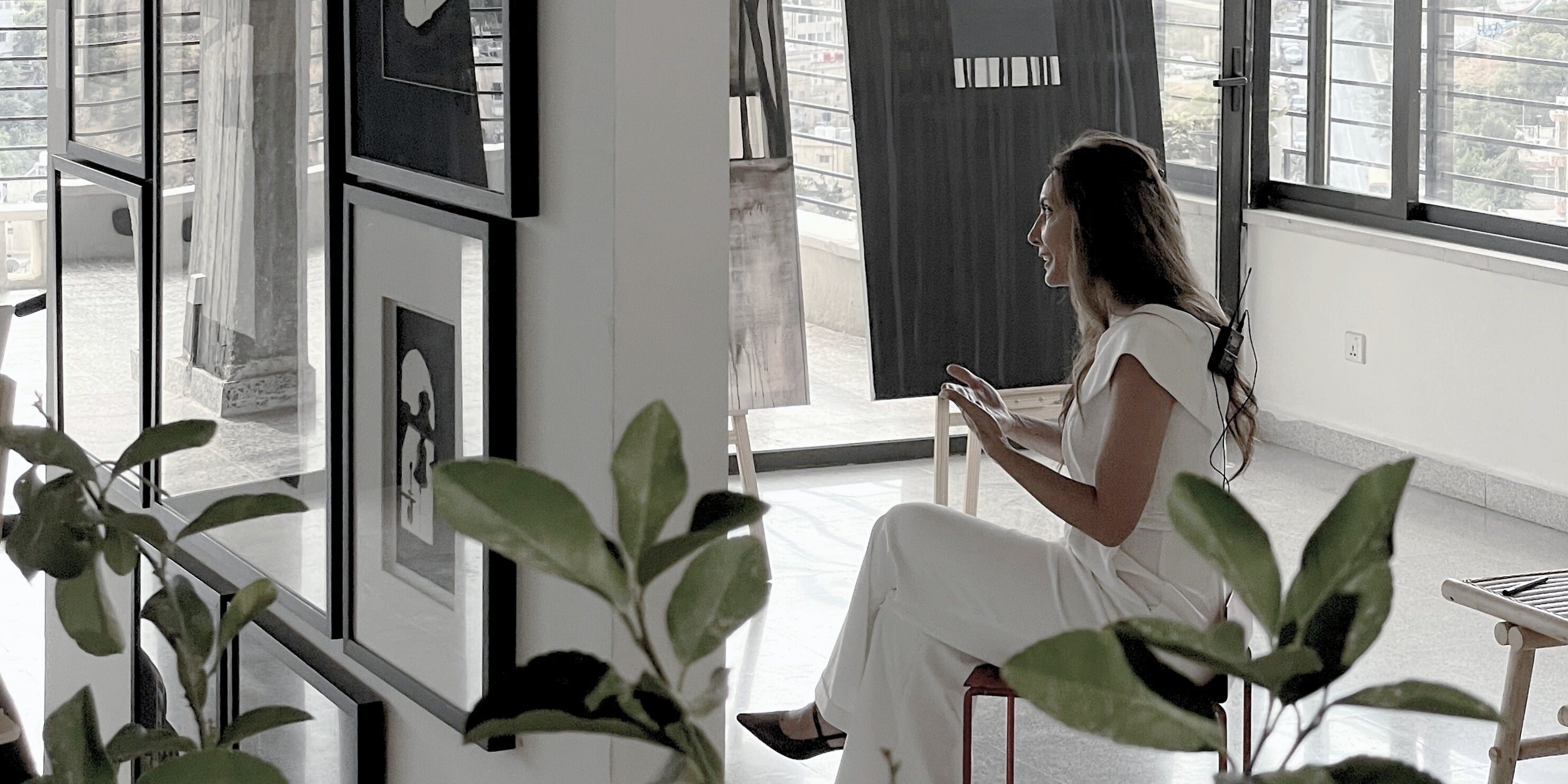 Rita Rawashdeh seated in a sunlit studio surrounded by framed artwork, talking with city views in the background.